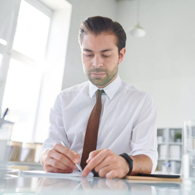 Young confident lawyer making working notes on paper while sitting by desk in office